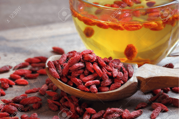 Gojibeeren in einem Holzlöffel mit einem Glas Gojibeeren Tee im Hintergrund